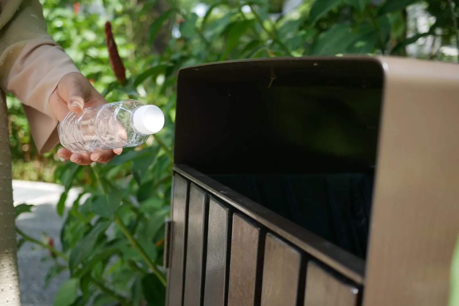 Close-up of a person dropping a plastic bottle into a recycling bin surrounded by greenery.
